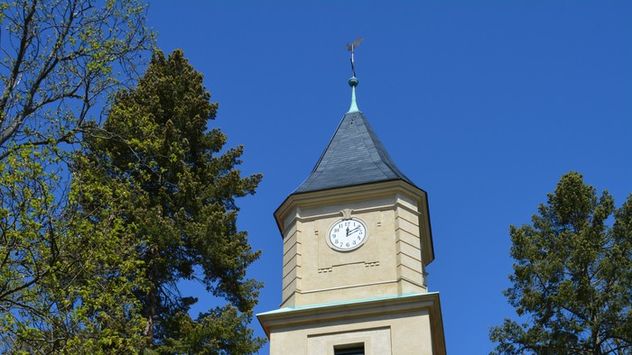 Der Kirchturm mit Uhr und spitzem Dach vor blauem Himmel.