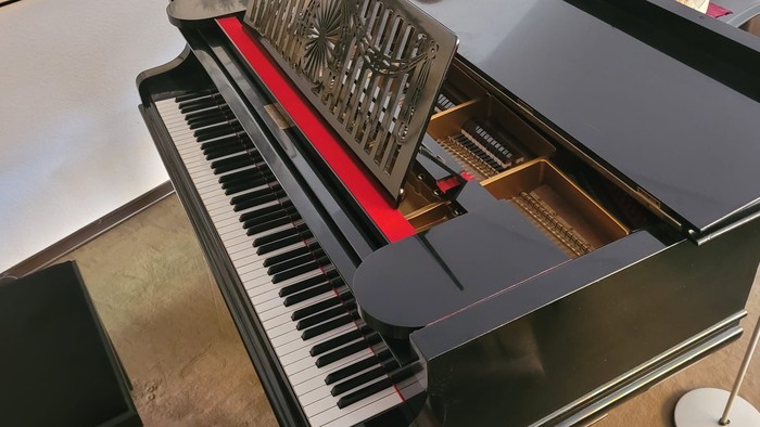 Modernes aufrechtes Klavier mit geöffnetem Deckel, das die komplexe innere Mechanik und ein rot akzentuiertes Resonanzboden zeigt.