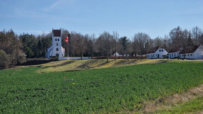 En hvid kirke med tårn og flag på en grøn åben mark med huse i baggrunden