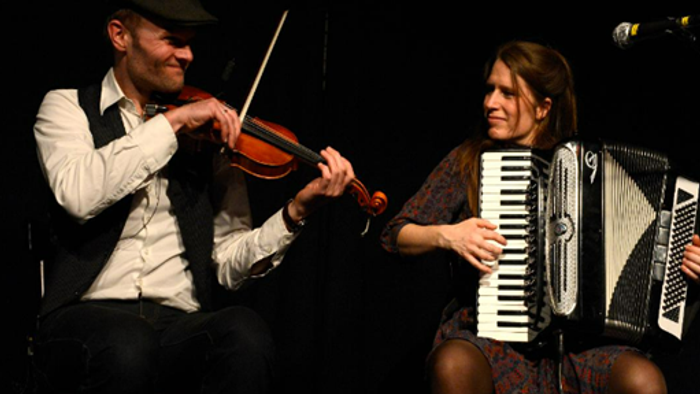 To musicere og en kvinde spiller violin og akkordeon på scenen