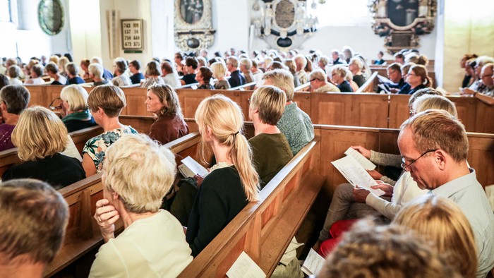 En kirke med mange tilstedeværende i bænkene, der ser ud til at deltage i en gudstjeneste.