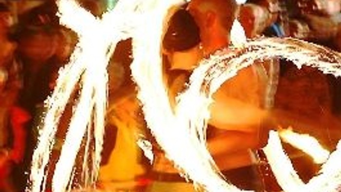 Eine Person führt mit Feuerstäben eine Show vor einem Publikum auf.
