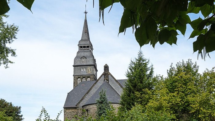 Ein Kirchengebäude mit hohem Turm und Uhr, umgeben von Bäumen und Büschen.
