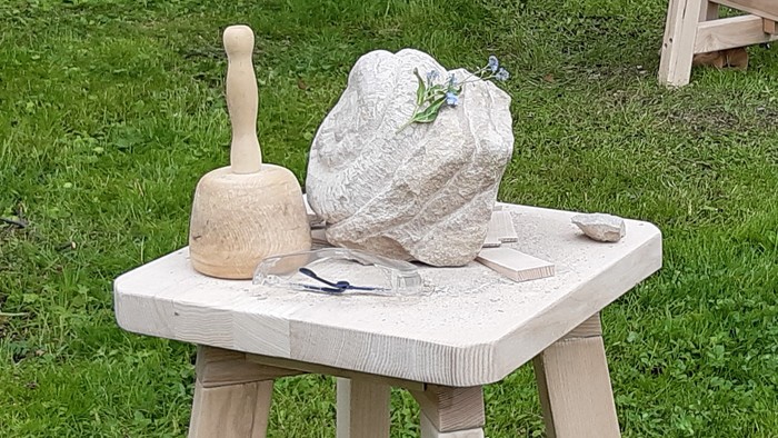 Auf einem Holztisch im Freien stehen ein großer Stein, ein Hammer und andere Werkzeuge.