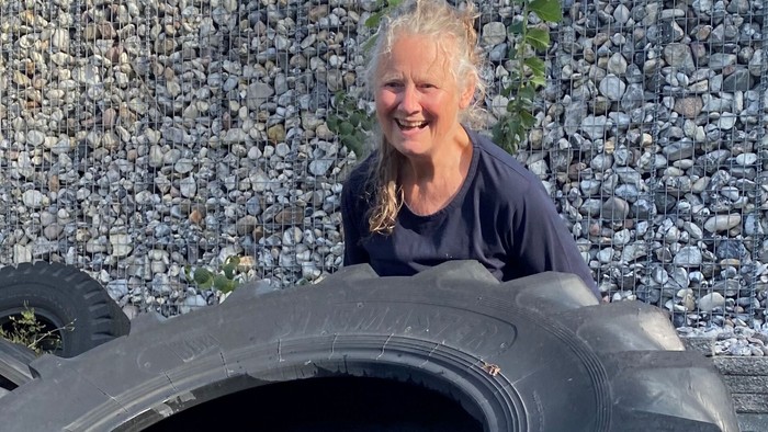 En glad person, der smiler, mens de sidder inde i et stort traktordæk foran en stenet væg.