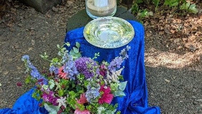 Ein ruhiger Outdoor-Altar mit Blumen, einer brennenden Kerze und einem offenen Buch auf blauem Tuch.