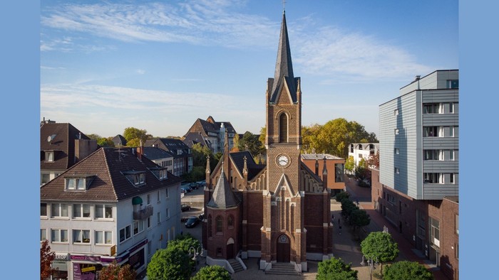 Luftaufnahme einer historischen Kirche, umgeben von modernen und traditionellen europäischen Gebäuden unter klarem Himmel.