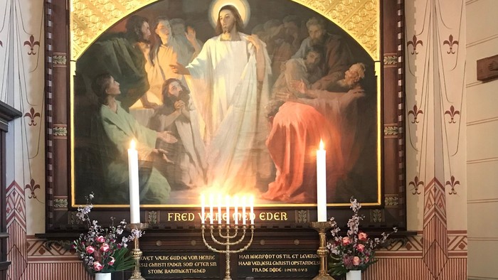 Religiøst alter med et vægmaleri af Jesus omgivet af figurer, lys og indskrevne plaketter.