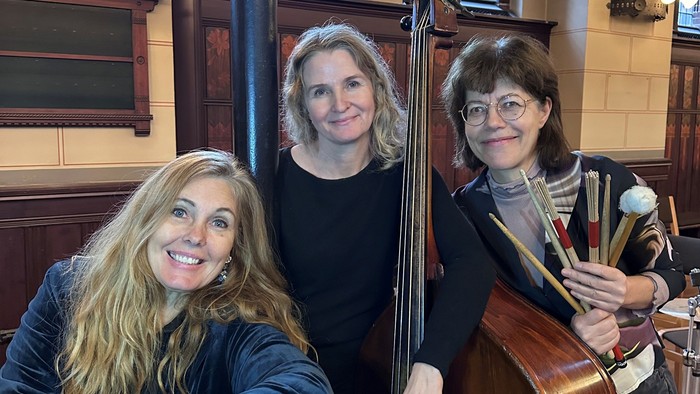 Tre musikere poser med strygeinstrumenter i en stor, historisk indendørsscene.