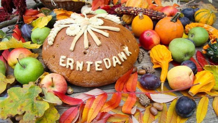 Ein herbstliches Arrangement mit einem Brot, das "Erntedank" lautet, umgeben von Früchten und bunten Blättern.