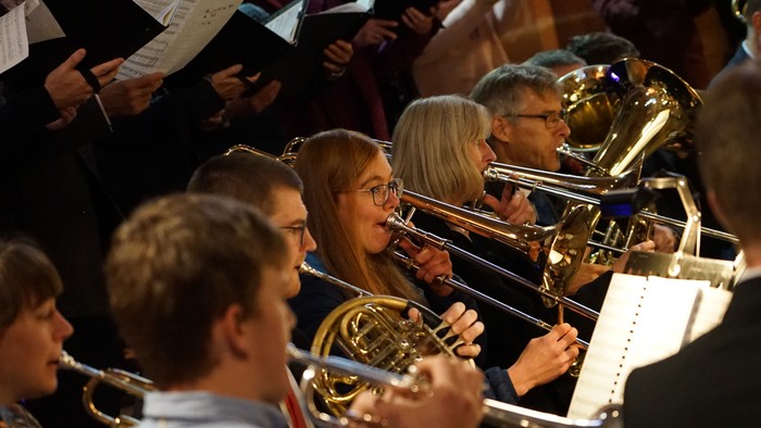 Ein Bläserensemble mit Trompeten, Posaunen und Hörnern spielt in einem Konzert. Ein Chor singt im Hintergrund.
