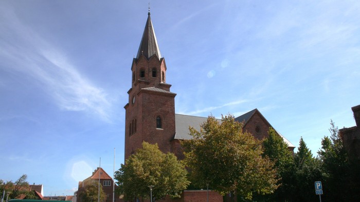 En murstenskirke med et højt spir står under en klar blå himmel omgivet af træer.