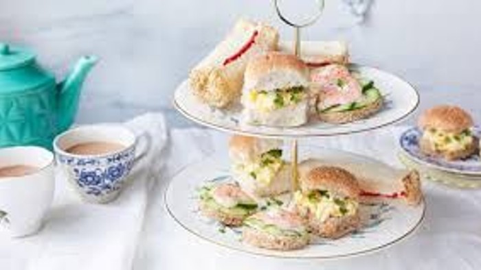 A tiered tray with sandwiches and a teapot, alongside cups and saucers on a marble surface