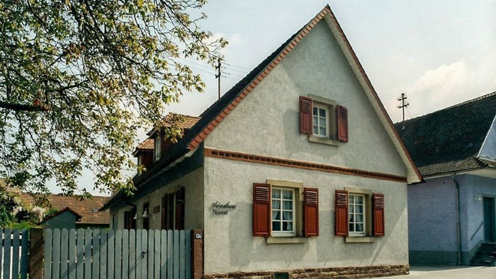 Das Bild zeigt ein charmantes, kleines Haus mit rotem Dach und weißen Fensterläden an einer Kopfsteinpflasterstraße.