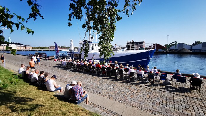 En gruppe af mennesker sidder på græs og bord ved vandet, hvor der er både store og små både i nærheden.