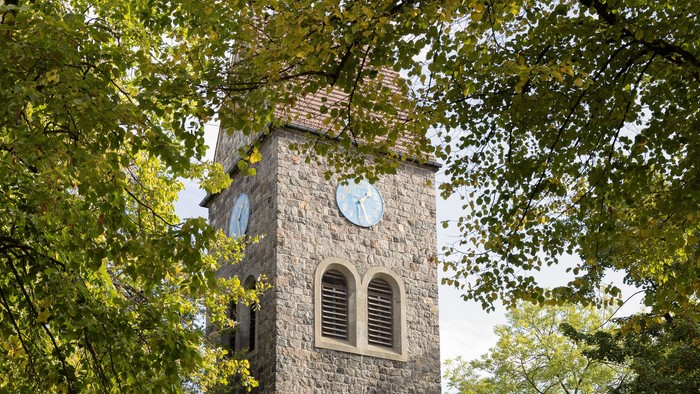 Der Kirchturm aus Stein mit Uhr und Fenster steht in einem Park.