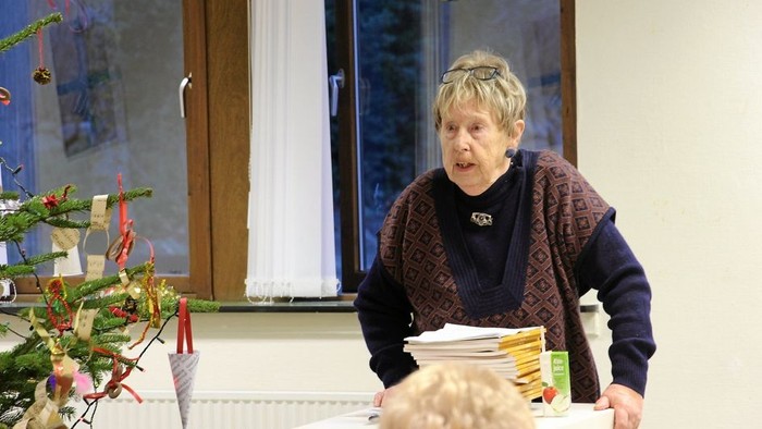 En kvinde står og underviser eller præsenterer for et barn, mens hun holder nogle papirer og står nær et pyntet juletræ.