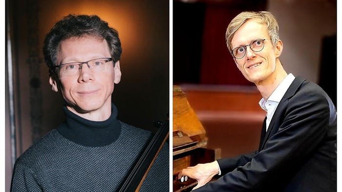 Zwei Männer spielen Klavier, einer trägt Brille und Pullover, der andere Anzug und Krawatte