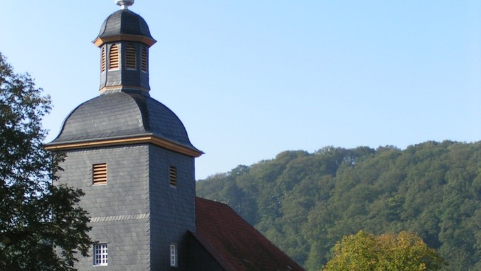 Kirche mit hohem Kirchturm und Kreuz, umgeben von Bäumen und Häusern.