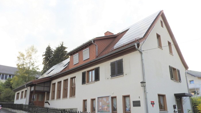 Zweistöckiges Haus mit rotem Dach und weißen Wänden, schwarzem Zaun und Solarpaneelen auf dem Dach.
