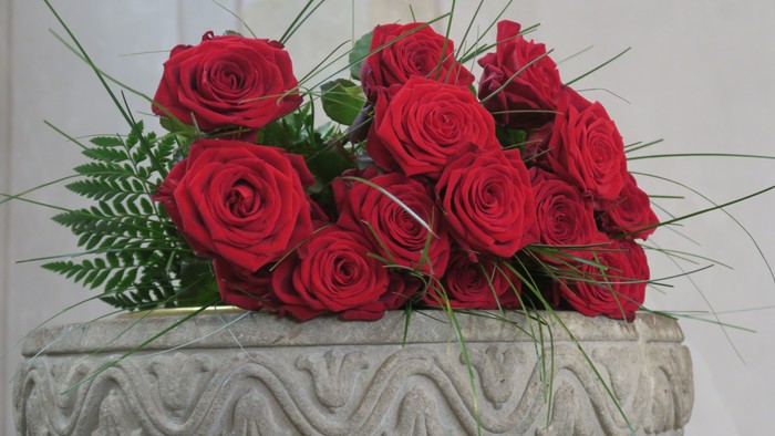 Man sieht den oberen Teil des Taufsteines in der Johanneskirche, er ist aus Sandstein und verziert, darauf liegen dekorativ einige rote Rosen.