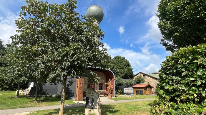 Man sieht das Gebäude des Knappenvereins, im Vordergrund steht ein Gedenkstein, im Hintergrund sieht man einen Kugelwasserturm.