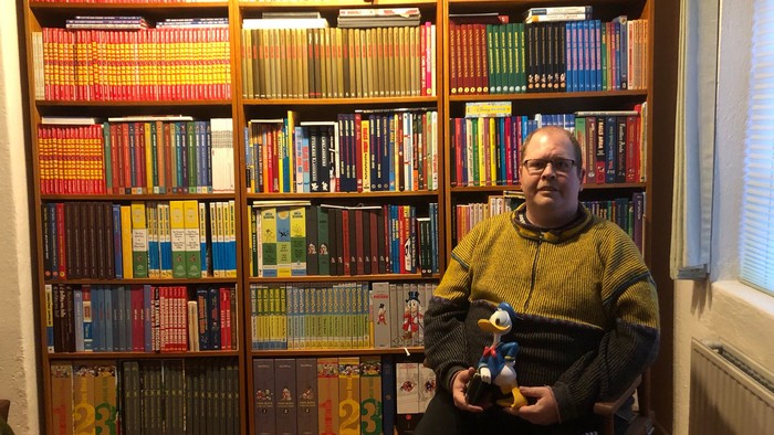 billede af foredragsholder foran sin bog reol og en figur af Anders And i hånden