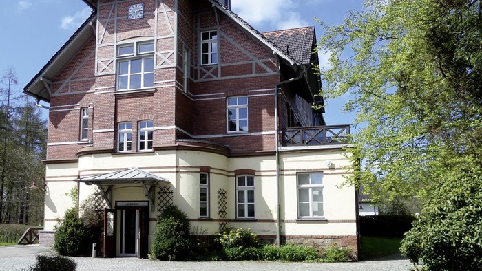 Das Bild zeigt ein zweistöckiges Holzhaus mit weißer Fassade und braunen Akzenten, umgeben von üppigem Grün.