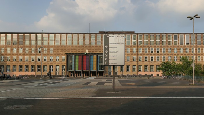 Grosses mehrstöckiges Gebäude mit farbenfrohem Wandgemälde an der Fassade, umgeben von Bäumen und Laternenpfählen.