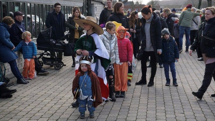En livlig udendørs sammenkomst med børn iført farverige kostumer og voksne, der spadserer rundt i en festlig atmosfære.