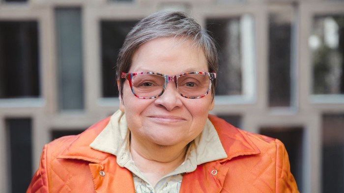 Eine Frau mit kurzem grauen Haar und bunten Brillengläsern lächelt vor einem Gebäude.