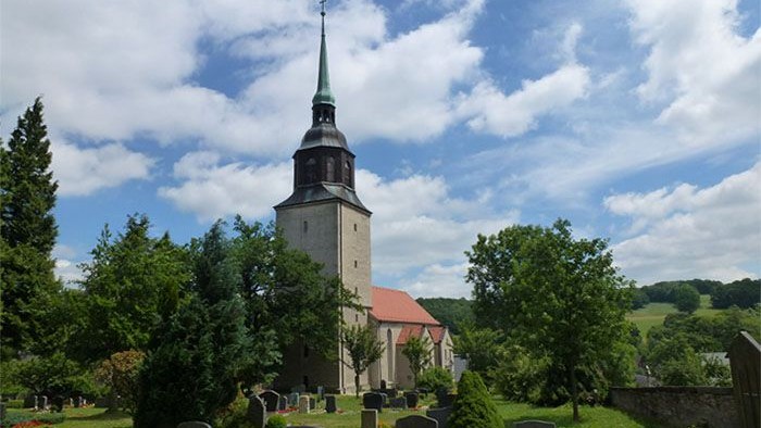 Kirchturm mit spitzem Dach und rotem Dach, umgeben von Bäumen und Gräbern