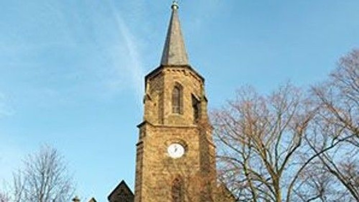 Ein hoher Kirchturm mit Uhr und spitzem Dach vor blauem Himmel und Bäumen.