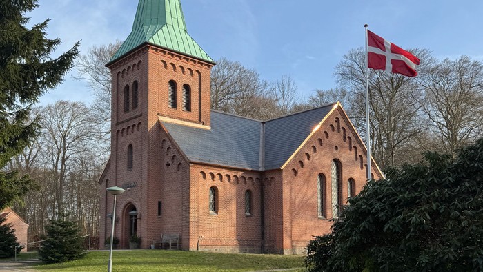 En rødstenskirke med et grønt spir og et dansk flag, der vajer ved siden af på en klar dag.