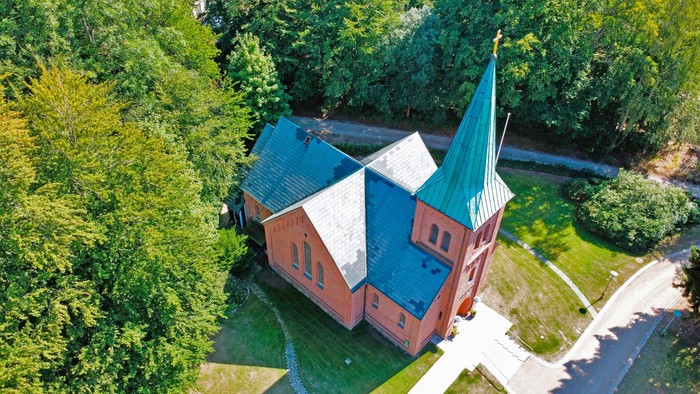 Luftfoto af en rødstenskirke med et grønt spir omgivet af en frodig, grøn skov.