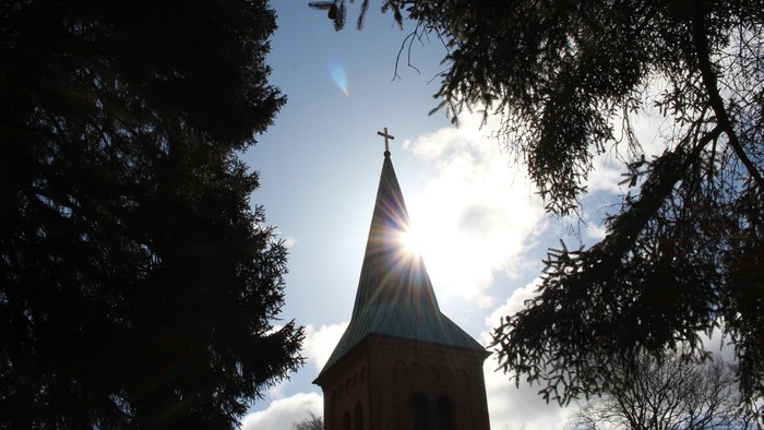 En kirke med spire og kors, omgivet af træer og solskin.