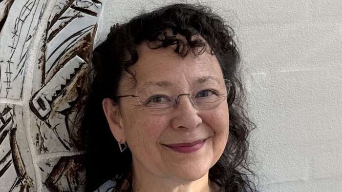 En kvinde med briller og halskæde står foran en mur med et mønster.