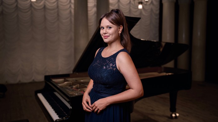 Junges Mädchen in blauem Kleid steht vor einem schwarzen Klavier.