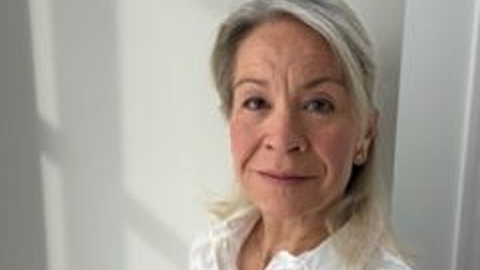 Eine Frau mit grauen Haaren lächelt sanft in einer weißen Bluse mit Muster in der Nähe eines Fensters.
