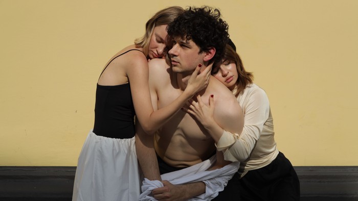 Drei Schauspieler führen auf der Bühne eine intime, emotional aufgeladene Szene mit körperlicher Nähe und dramatischen Ausdrucksformen auf.