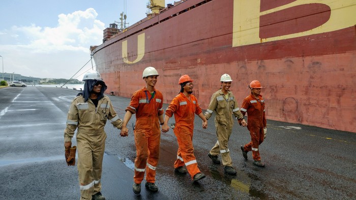 Arbeiter in orangen Anzügen gehen in der Nähe eines großen Schiffes.
