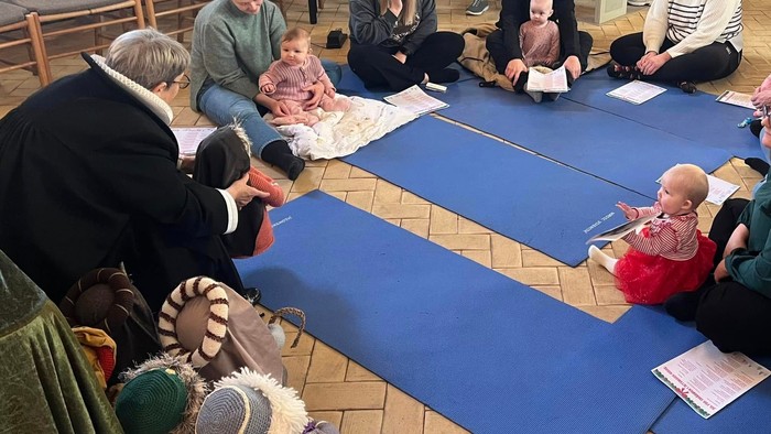 En gruppe af voksne og børn sidder på gulvmatte i et rum med piano.