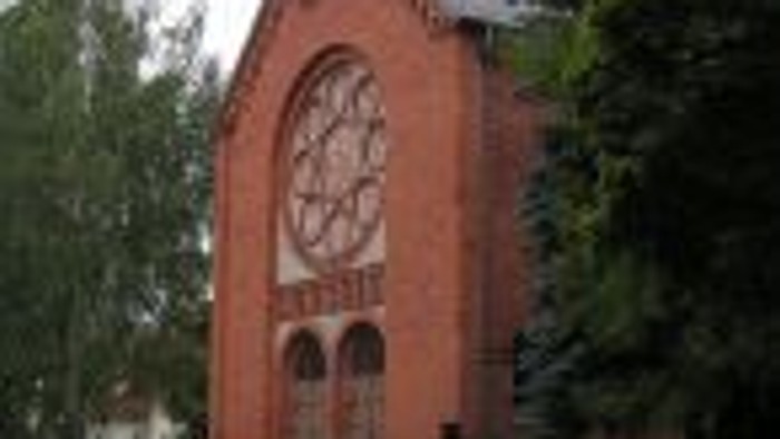 Eine rote Backsteinkirche mit Bogenfenstern und einem hohen Kirchturm, umgeben von üppigen grünen Bäumen.