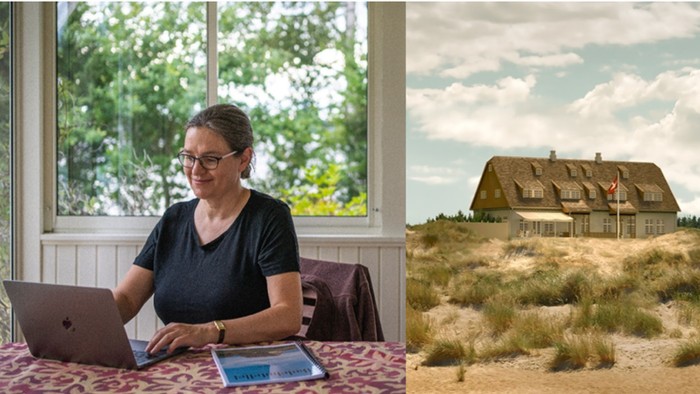 En kvinde arbejder på sin laptop i en lys og rummelig stue med udsigt til en træhus i en naturlig omgivelse.