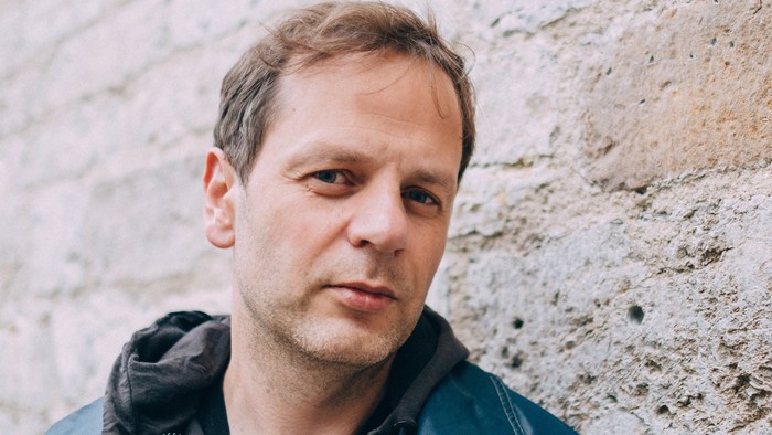 Ein Mann in einer dunklen Jacke lehnt mit nachdenklichem Gesichtsausdruck an einer strukturierten Steinmauer.