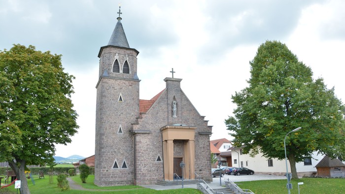 Kleine Steinkirche mit Glockenturm und Kreuz, umgeben von Bäumen und Gras.