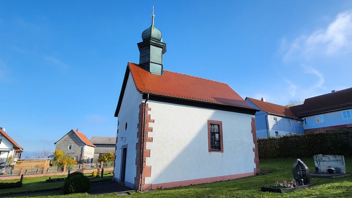Weißes Kirchegebäude mit roter Ziegeldach und kleiner Kuppel, umgeben von grünem Gras und anderen Häusern