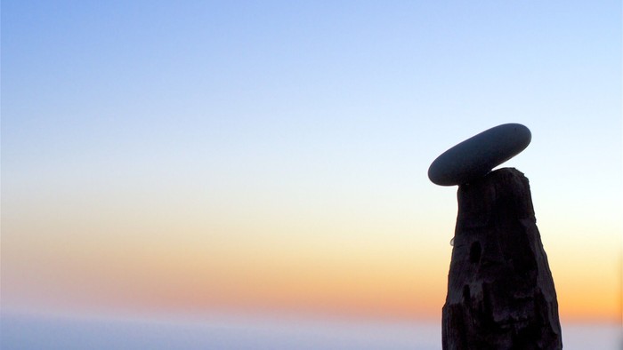 Ein Kieselstein liegt auf einem angespitzen Baumpfahl. Im Hintergrund sieht man das Meer im Sonnenuntergang