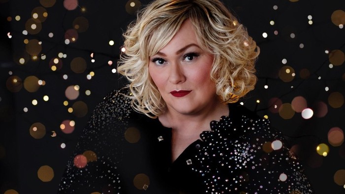 Blond kvinde med krøllet hår iført en sort paljet-top, der poser mod en mørk baggrund med bokeh-lys.