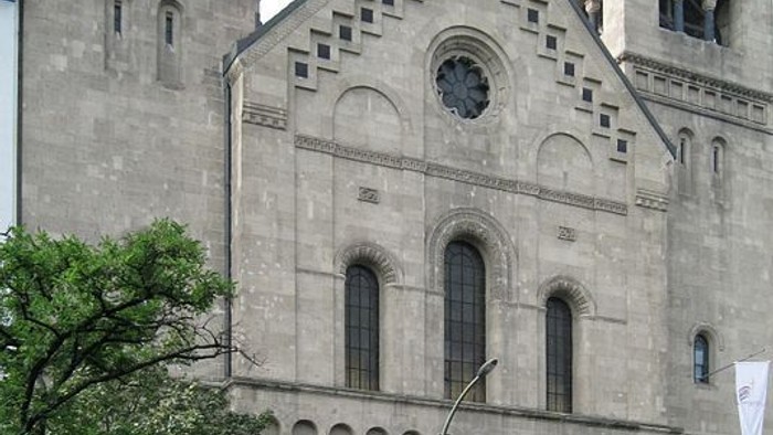 Großes, altes Gebäude mit zwei Türmen und Bogenfenstern, daneben Parkplatz mit rotem Auto